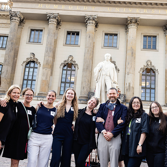 Eine Gruppe Humboltdianer*innen vor dem HU-Hauptgebäude Arm in Arm in die Kamera schauend