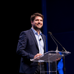 Matthias König steht vor einem Rednerpult mit Mikrofon und lächelt ins Publikum. Er trägt ein hellblaues Hemd und ein dunkelblaues Sakko.