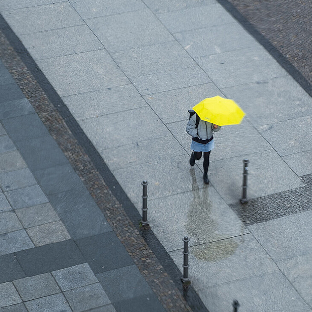 Eine Person spaziert auf einer breiten Straße mit einem knallgelben Regenschirm in der Hand. Das Foto ist von oben aufgenommen.
