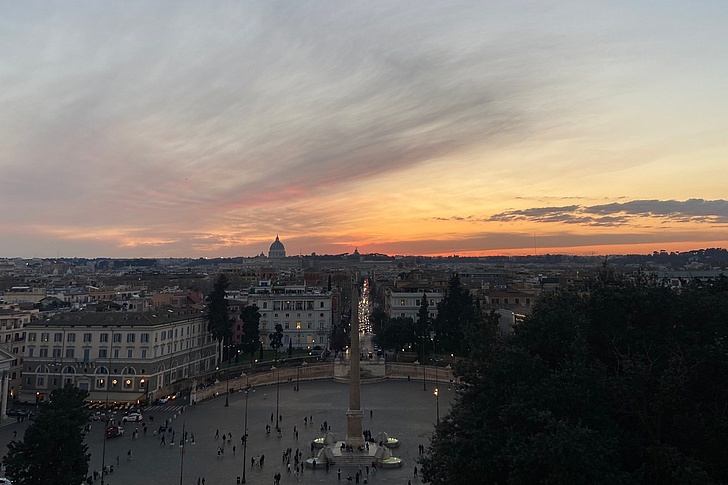 Sonnenuntergang über Piazza del Popolo in Rom: Orangefarbener Himmel über Kuppelkirchen, Obelisk, Springbrunnen und Menschenmengen mit Palmen und Gebäuden.