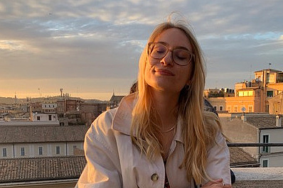 Blonde Frau mit Brille und langen Haaren in beigem Mantel lächelt genüsslich mit geschlossenen Augen bei Sonnenuntergang über der orange erleuchteten Skyline von Rom mit Dächern und Türmen.