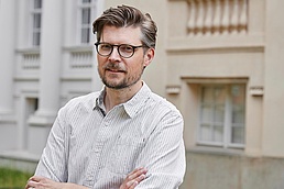 Prof. Dr. Tobias Krüger steht mit verschränkten Armen vor einem Gebäude der Humboldt-Universität- Er trägt ein hell-gestreiftes Hemd.