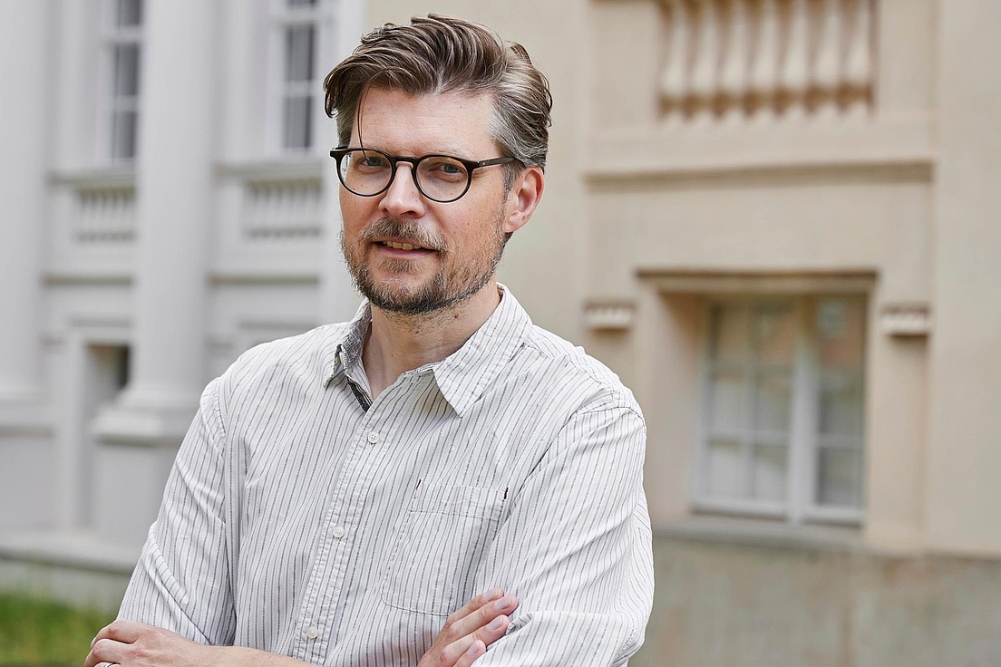 Prof. Dr. Tobias Krüger steht mit verschränkten Armen vor einem Gebäude der Humboldt-Universität- Er trägt ein hell-gestreiftes Hemd.