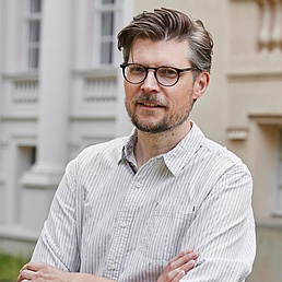Prof. Dr. Tobias Krüger steht mit verschränkten Armen vor einem Gebäude der Humboldt-Universität- Er trägt ein hell-gestreiftes Hemd.