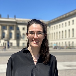 Person with long dark hair wearing a black shirt standing in front of a historic building with columns and many windows