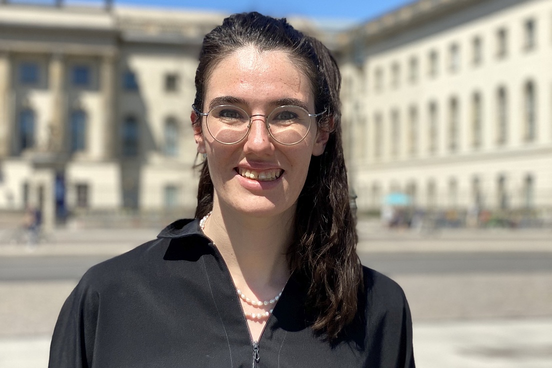 Person mit dunklem, langem Haar und schwarzem Hemd steht vor einem historischen Gebäude mit Säulen und vielen Fenstern