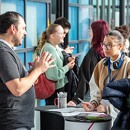 Im Bild sind verschiedene Personen in kleinen Gruppen zu sehen, die miteinander reden und diskutieren. Im Vordergrund steht ein Mann in einem grauen Shirt hinter einem weißen Stehtisch und hat seine Hände in der Luft, um etwas zu verdeutlichen. Vor dem Tisch hören zwei weibliche Personen in Freizeitkleiung zu