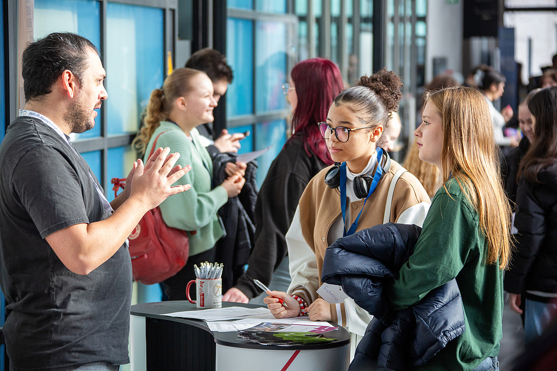 Im Bild sind verschiedene Personen in kleinen Gruppen zu sehen, die miteinander reden und diskutieren. Im Vordergrund steht ein Mann in einem grauen Shirt hinter einem weißen Stehtisch und hat seine Hände in der Luft, um etwas zu verdeutlichen. Vor dem Tisch hören zwei weibliche Personen in Freizeitkleiung zu