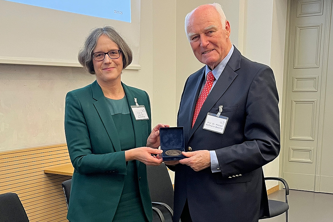 Michael Hoffmann-Becking trägt einen dunkelfarbenen Anzug, eine rot-weiß-gemusterte Krawatte und ein hellblaues Hemd. Neben ihm steht Julia Blumenthal, die ein grünes Kostüm trägt. Sie überreicht ihm die Humboldt-Universitäts-Medaille.