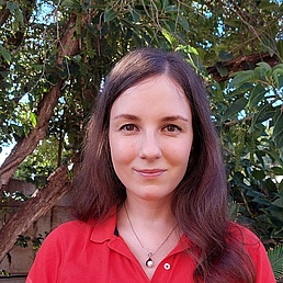 Woman with long brown hair wearing a red polo shirt and a necklace with a round pendant standing in front of a tree with green leaves.