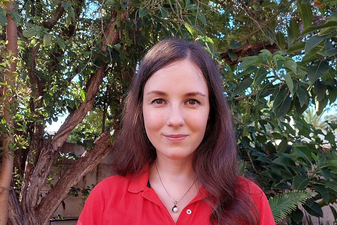 Frau mit langen braunen Haaren trägt ein rotes Poloshirt und eine Halskette mit rundem Anhänger vor einem Baum mit grünen Blättern.
