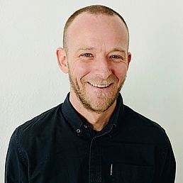 Julian Hamann lächelt in die Kamera und steht vor einer hellen Wand und trägt ein schwarzes Hemd
