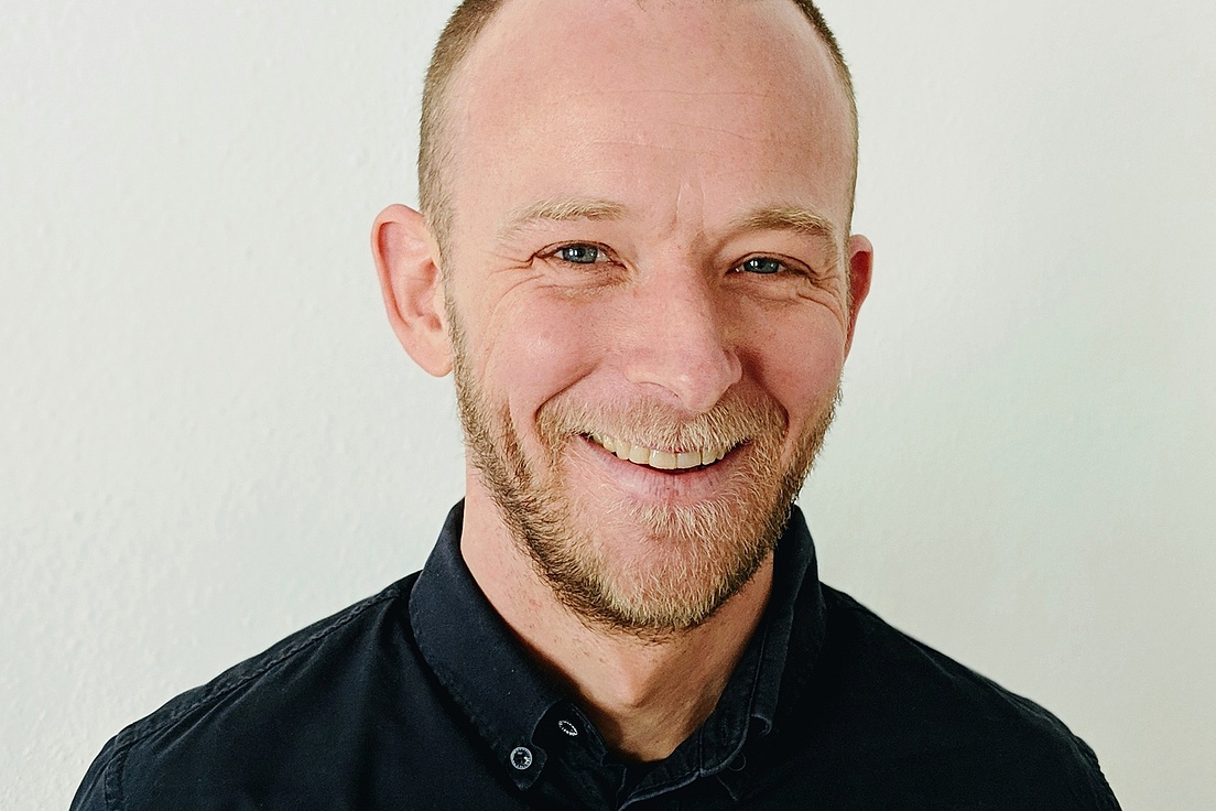 Julian Hamann lächelt in die Kamera und steht vor einer hellen Wand und trägt ein schwarzes Hemd