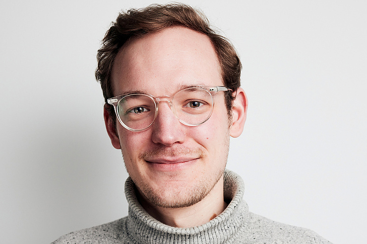Ein Mann mit Brille und grauem Rollkragenpullover lächelt vor weißem Hintergrund.