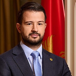 Jakov Milatović in Anzug und Krawatte mit Bart, vor einer Flagge und Vorhängen.