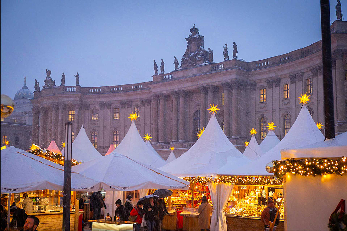 Beleuchtete Buden auf. dem Weihnachtsmarkt auf dem Bebelplatz. Sie haben ein Zeltdach und ein beleuchteter Stern ist auf  der Zeltspitze zu sehen. Im Hintergrund ist die Juristische Fakultät der HU zu sehen.