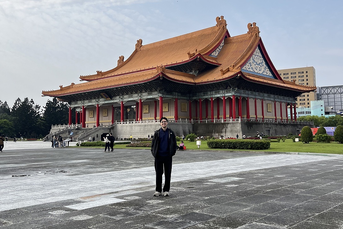 Person steht vor einem großen traditionellen asiatischen Gebäude mit geschwungenem Dach und roten Säulen auf einem weitläufigen gepflasterten Platz