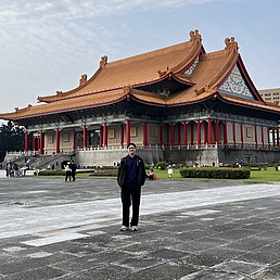 Person steht vor einem großen traditionellen asiatischen Gebäude mit geschwungenem Dach und roten Säulen auf einem weitläufigen gepflasterten Platz