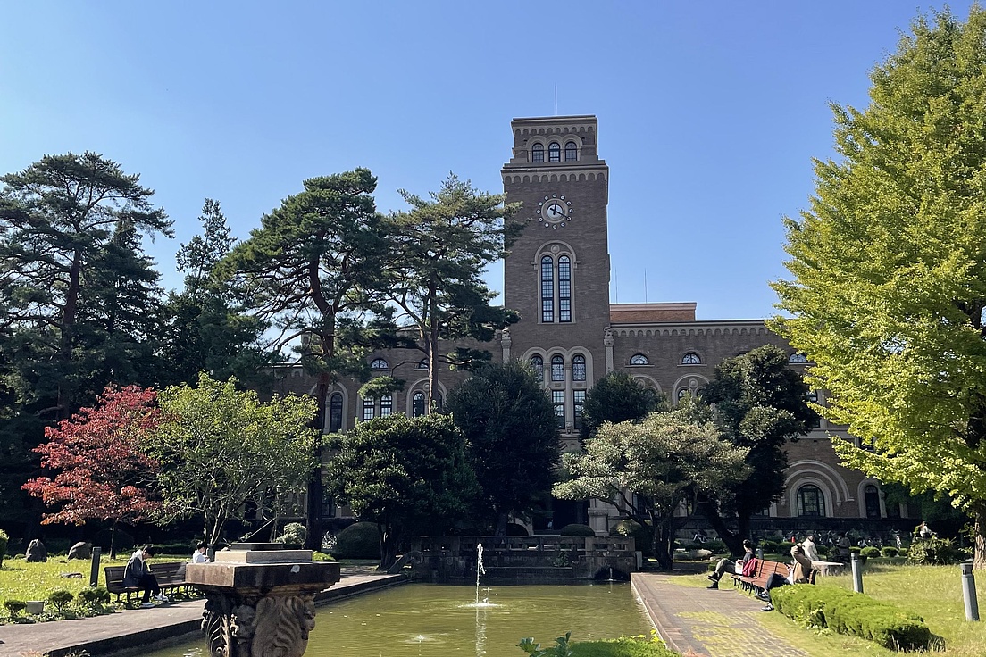 Historisches Gebäude mit Uhrenturm hinter einem Teich und Bänken in einem Park mit Bäumen