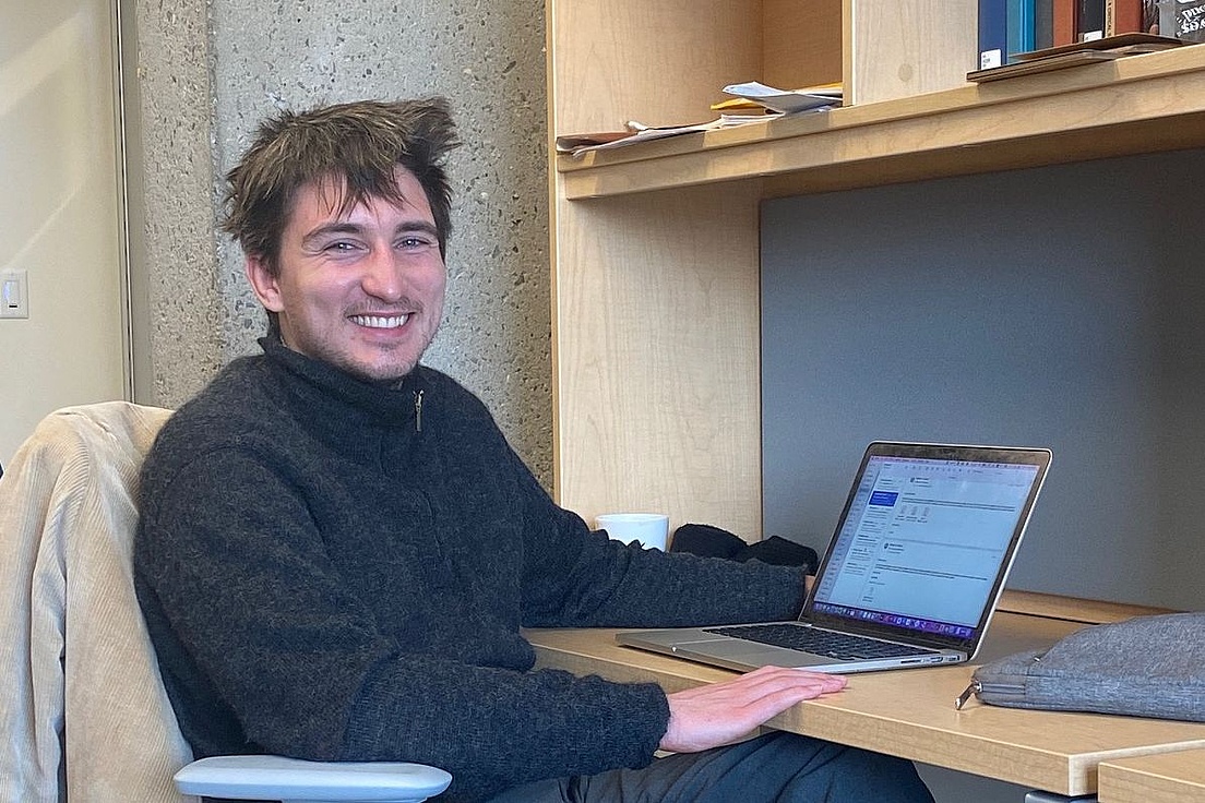 Junger Mann mit zerzaustem dunklem Haar und breitem Grinsen in grauem Rollkragenpullover sitzt an einem Holztisch mit Laptop, Kaffeetasse und Büchern in einer modernen Bibliothek.