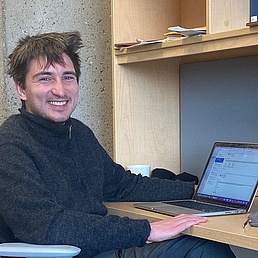 Junger Mann mit zerzaustem dunklem Haar und breitem Grinsen in grauem Rollkragenpullover sitzt an einem Holztisch mit Laptop, Kaffeetasse und Büchern in einer modernen Bibliothek.