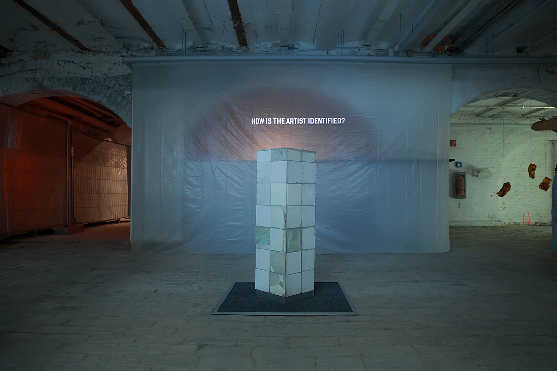 Ausstellung in der Kleinen Humboldt Galerie Eine Installation in einem kargen, industriell wirkenden Raum mit Betonboden und sichtbaren Rohren an der Decke. Im Zentrum steht ein zylindrischer Sockel aus weißen Fliesen, der auf einer dunklen Matte ruht. Hinter dem Sockel ist eine große, durchscheinende Plastikplane aufgespannt, auf der die Frage „HOW IS THE ARTIST IDENTIFIED?“ in weißer Schrift projiziert wird