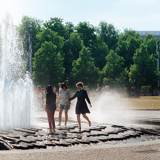 Drei Personen, die sich an einer großen Wasserfontäne abkühlen.