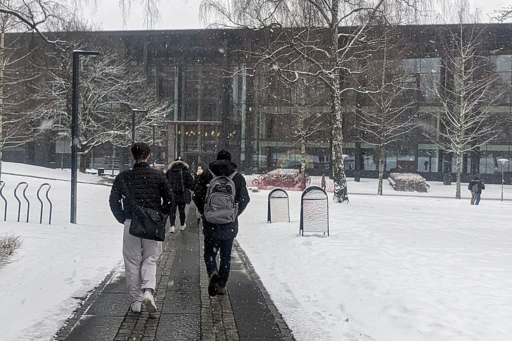 Menschen mit Winterkleidung und Rucksäcken gehen auf einem schneebedeckten Weg zu einem modernen Gebäude mit großen Fenstern und kahlen Bäumen auf beiden Seiten.