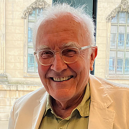 Bernd Hennigsen trägt ein helles Hemd und darüber eine helle Jacke und trägt eine randlose Brille. Er lächelt in die Kamera und steht vor einem Fenster. Im Hintergrund ist ein altes Gebäude zu sehen.