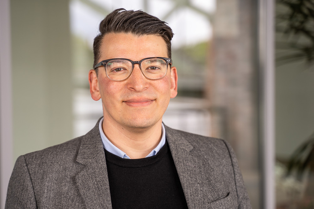 Christopher Degelmann mit Blazer, schawrzem Pullover und Brille.  Im Hintergrund sieht man ein Fenster.