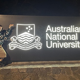 Person mit gestreiftem Pullover und Rock posiert neben beleuchtetem Schild der Australian National University mit Wappen und lateinischem Motto.