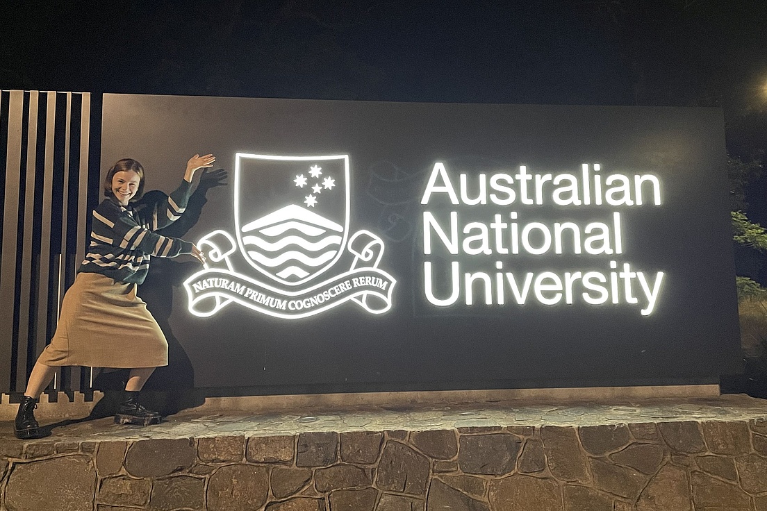 Person mit gestreiftem Pullover und Rock posiert neben beleuchtetem Schild der Australian National University mit Wappen und lateinischem Motto.
