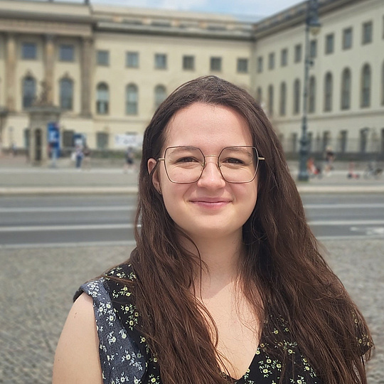 Frau mit langen, offenen Haaren und ärmellosem, gemustertem Oberteil steht vor einem historischen Gebäude mit Säulen und mehreren Fenstern.