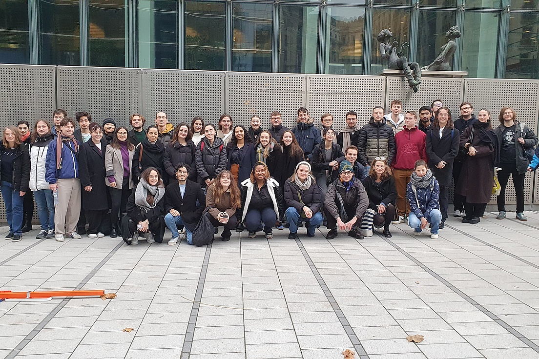 Around 50 people gather for a group photo in front of an urban building with a glass façade and sculpture.