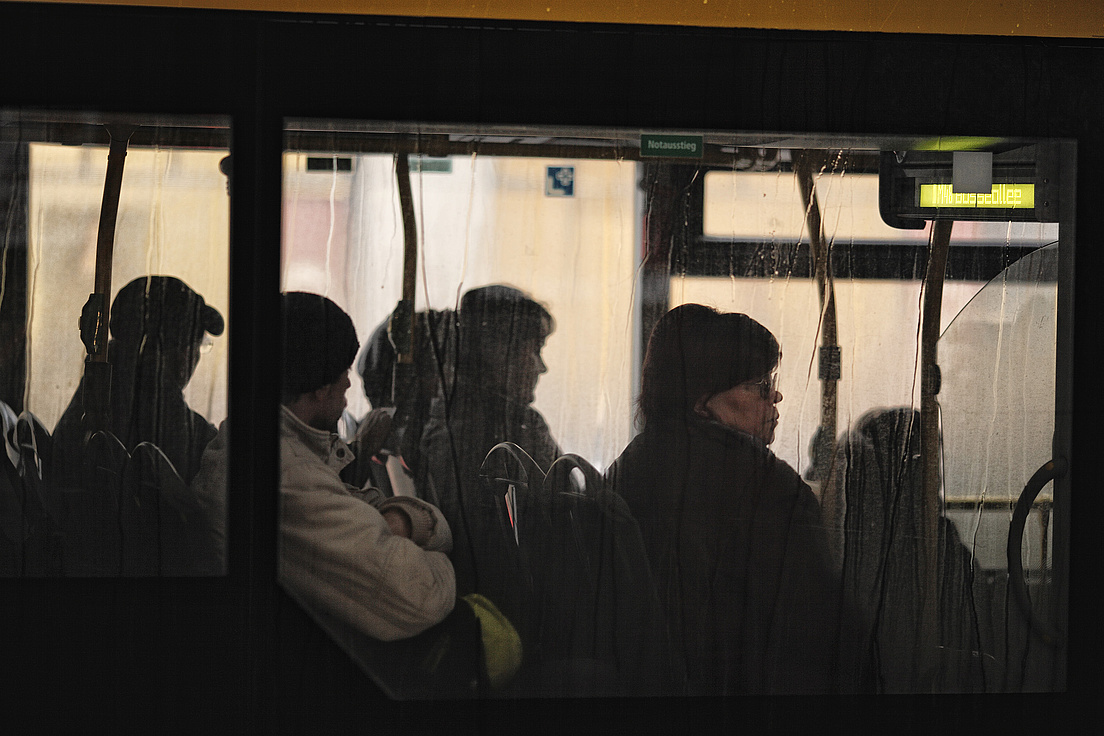 Das Bild zeigt eine Gruppe von Menschen, die in einem öffentlichen Verkehrsmittel, vermutlich einem Bus, stehen. Die Szene ist durch das Fenster des Busses fotografiert worden, wodurch Regentropfen auf der Scheibe sichtbar sind. Die Personen sind in dunkle Kleidung gehüllt.