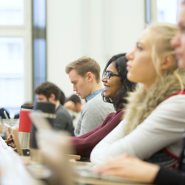 Ein Vorlesungssaal. Man sieht mehrere Studierende. Eine Person sitzt in der Mitte. Sie sticht besonders heraus, eine junge Frau lächelt und schaut zur Lehrkraft.