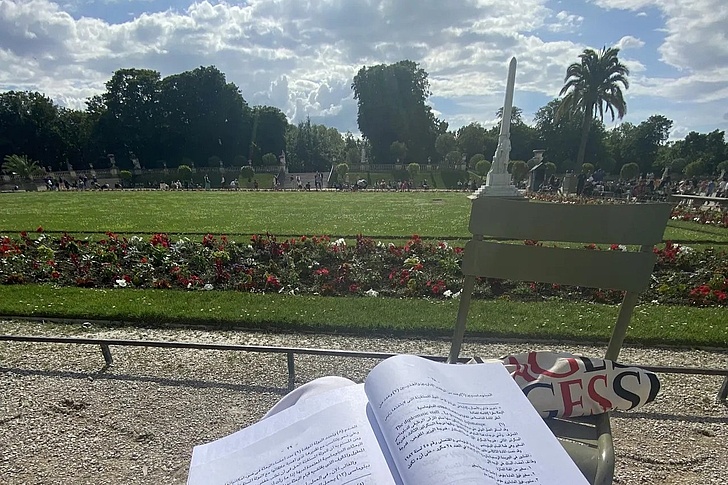 Blick über ein offenes Buch auf eine Parkwiese mit Blumenbeet, Stuhl und Bäumen im Hintergrund