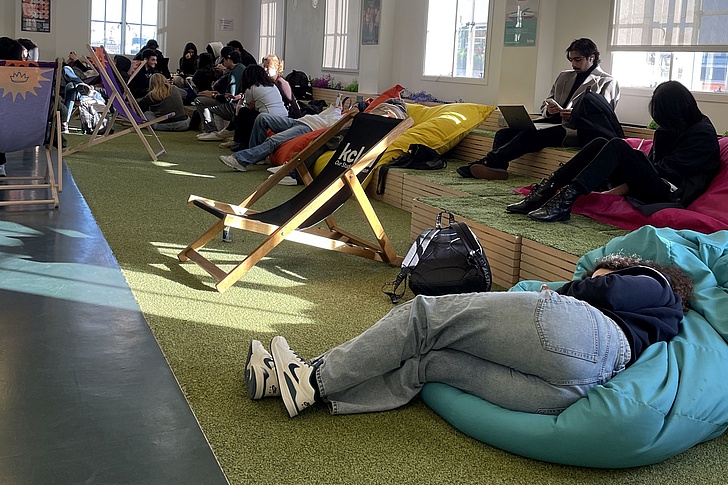 Mehrere Personen sitzen und liegen in einem hellen Raum auf Sitzsäcken, Liegestühlen und einer erhöhten Sitzfläche mit Kissen.
