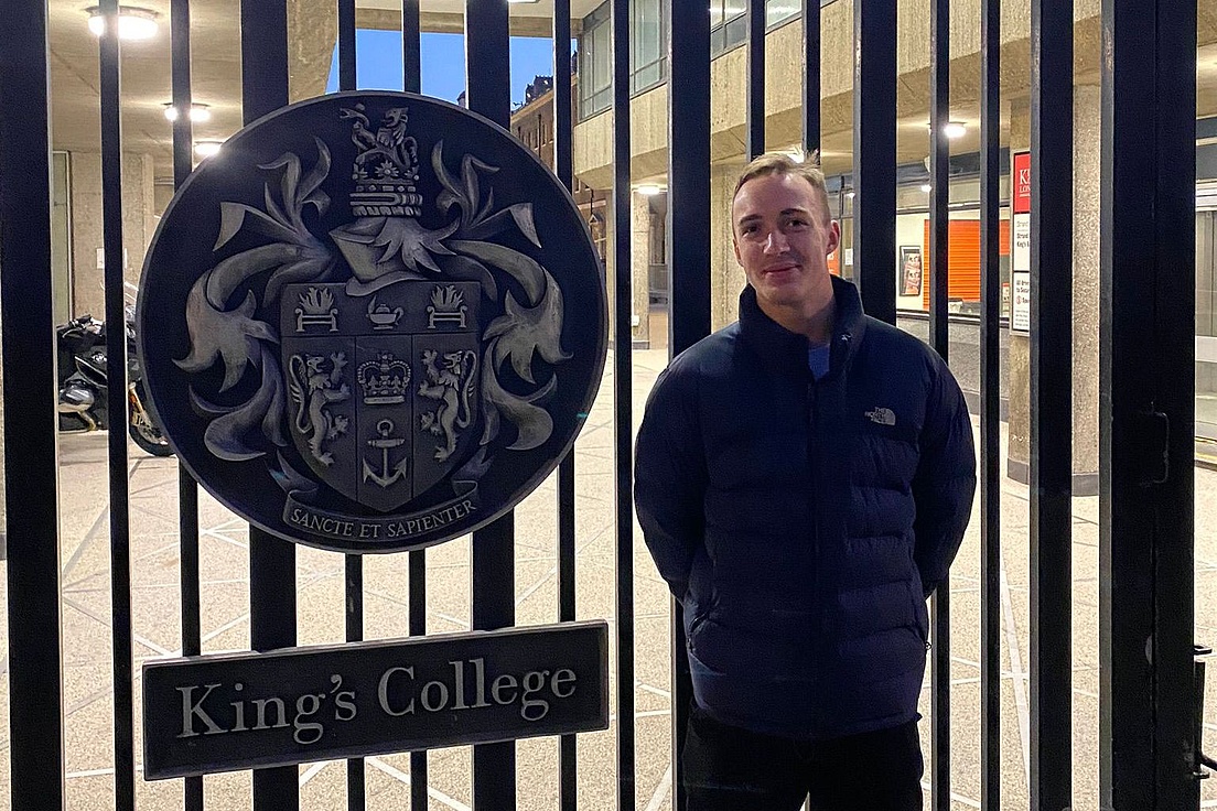 Person steht vor einem schwarzen Metalltor mit dem Wappen und Schild von King's College.