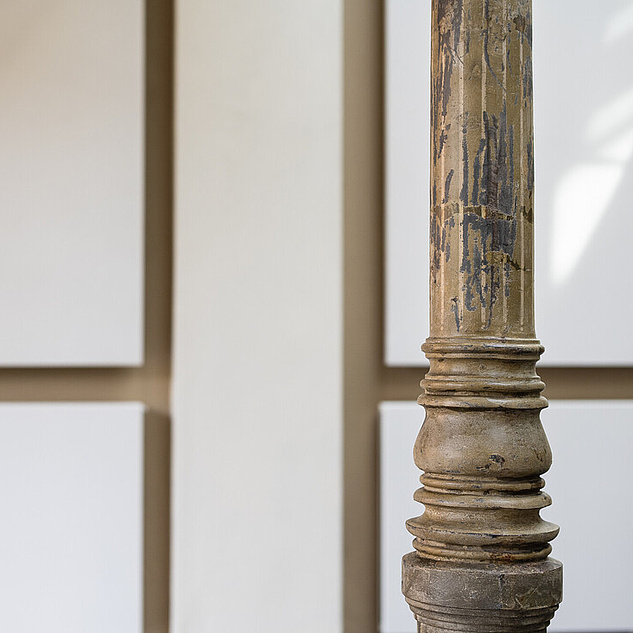 Campus Nord Eine Wand, die wie mit goldenen Linien leicht gekachelt aussieht. Davor eine alte schmale Säule, kunstvoll mit Rillen.