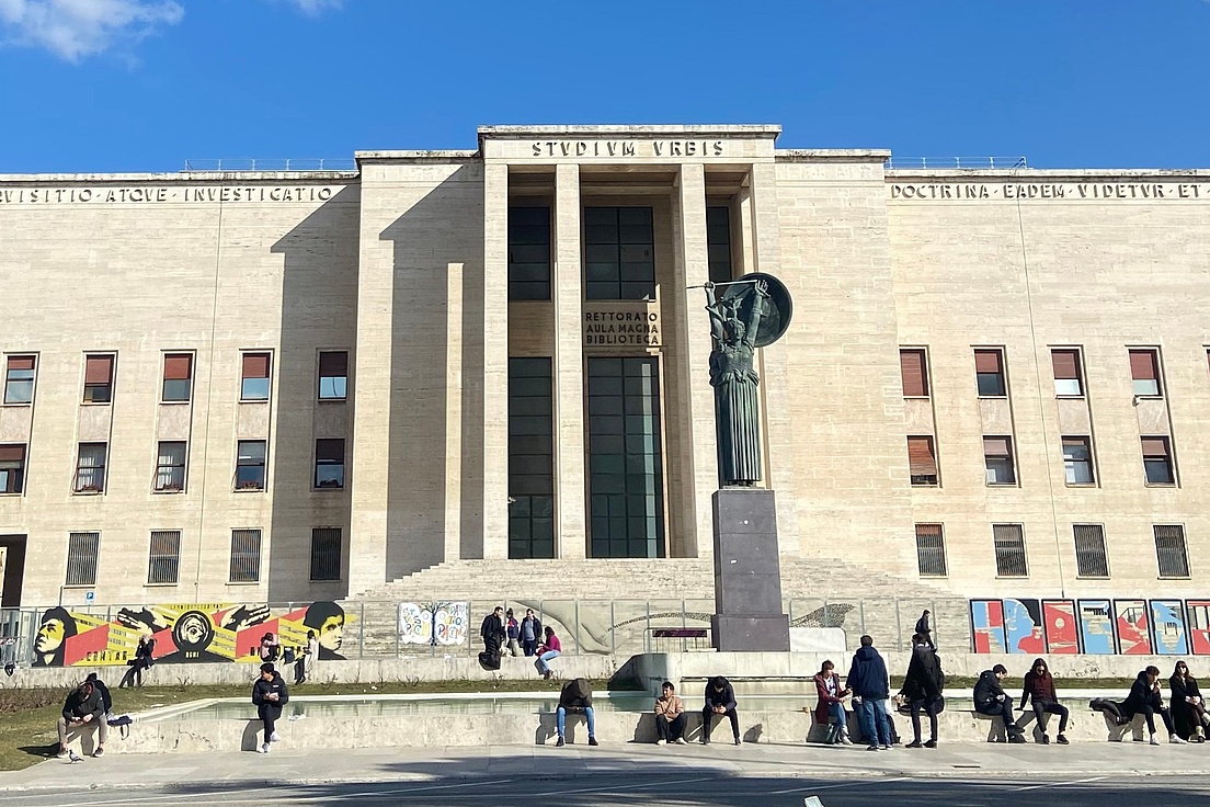 Fassade des Stadio di Domiziano in Rom: Beige Steinarchitektur mit zentraler Statue, bunten Protestplakaten und Menschengruppe davor an sonnigem Tag.