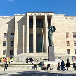 Fassade des Stadio di Domiziano in Rom: Beige Steinarchitektur mit zentraler Statue, bunten Protestplakaten und Menschengruppe davor an sonnigem Tag.