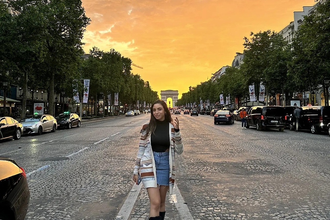 Frau mit Jeansrock und langem Cardigan steht auf der Straße vor dem Arc de Triomphe bei Sonnenuntergang
