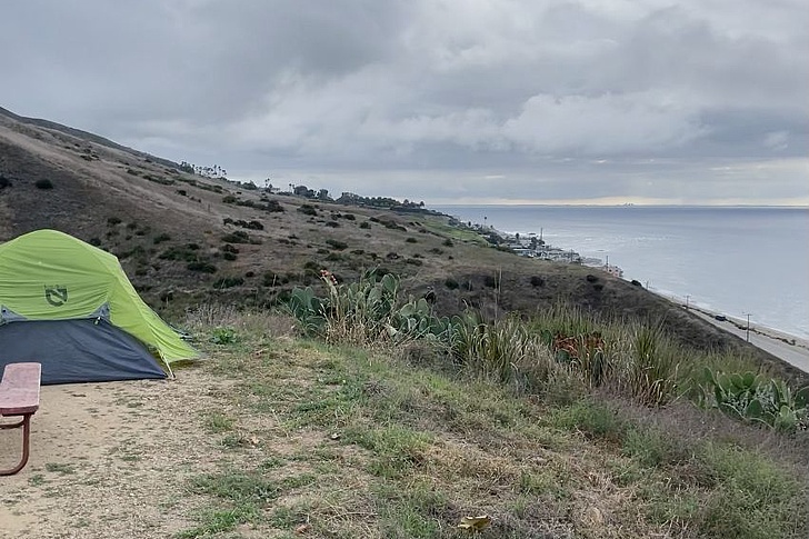 Grüne Campingzelt auf einem grasigen Hügel mit Blick auf das Meer unter bewölktem Himmel