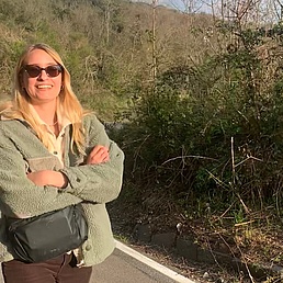 Lächelnde blonde Frau mit Sonnenbrille in grüner Felljacke und schwarzer Tasche lehnt mit verschränkten Armen an einer kurvigen Straßenböschung inmitten grüner Hügel bei sonnigem