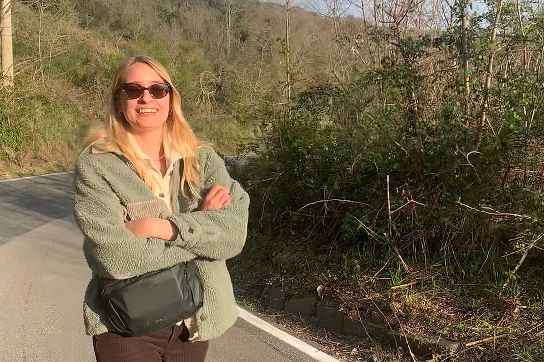 Lächelnde blonde Frau mit Sonnenbrille in grüner Felljacke und schwarzer Tasche lehnt mit verschränkten Armen an einer kurvigen Straßenböschung inmitten grüner Hügel bei sonnigem
