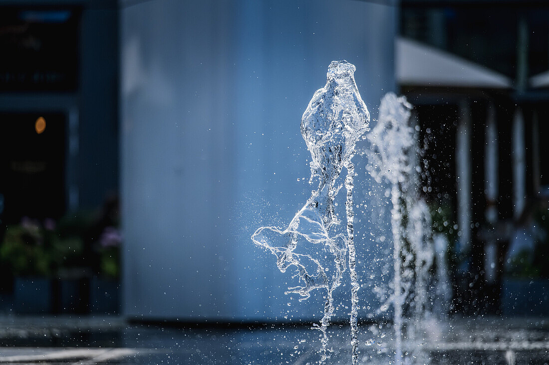 Das Bild zeigt eine Nahaufnahme eines Wassersprudlers, der in einem Brunnen oder einer Fontäne zu sehen ist. Im Hintergrund sind unscharfe Elemente einer städtischen Umgebung zu erkennen.