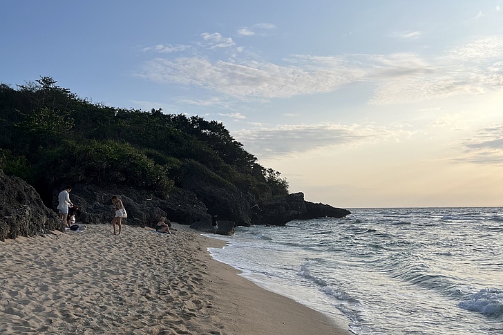 Strand mit Sand, Felsen und mehreren Personen am Wasser unter bewölktem Himmel