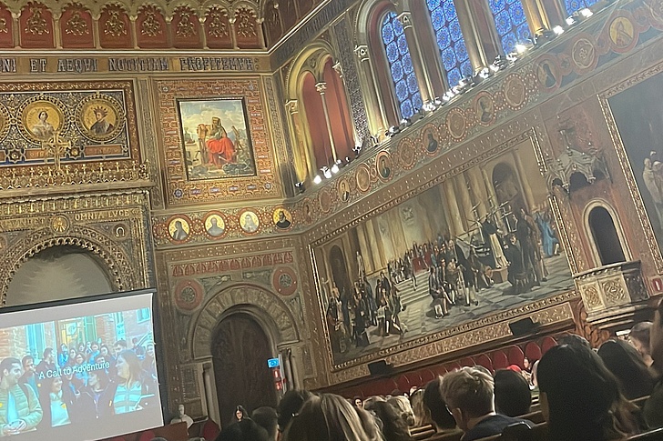 Großer Saal mit reich verzierten Wänden und hohen Fenstern, Publikum sitzt auf Holzbänken, links eine Leinwand mit einer Projektion.