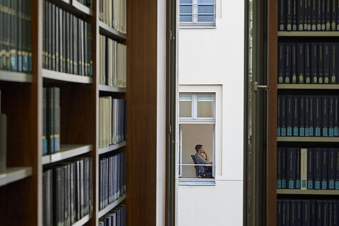 Eine Person sitzt auf einem Stuhl an einem Tisch. Das Foto ist so aufgenommen, dass die Person durch ein schmales Fenster fotografiert wurde. Man sieht sie nur von der Seite im gegenüberliegenden Gebäude. Im Vordergrund sind Bücherregale aus dunklem Holz zu sehen. 